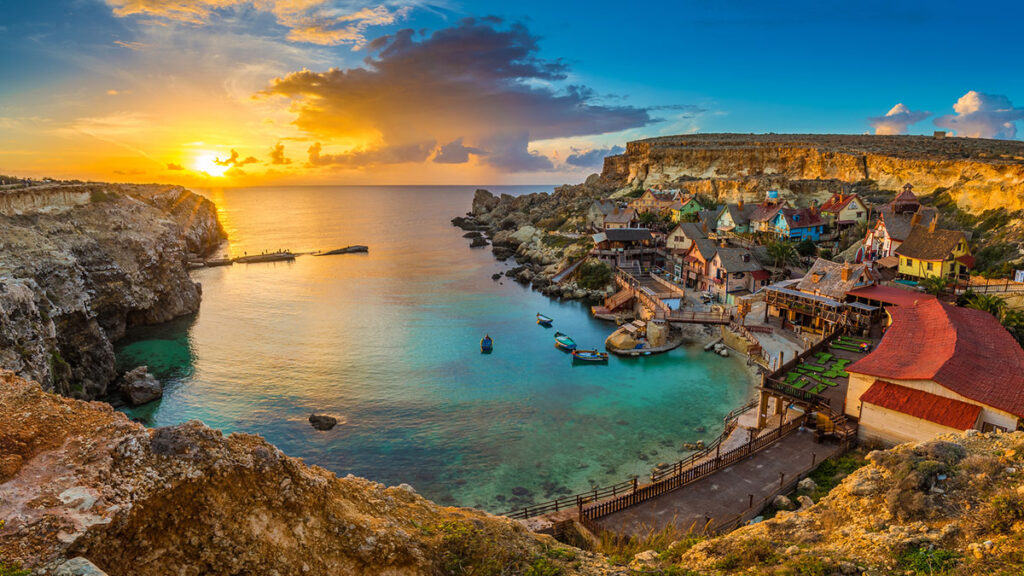 Panoramic skyline view of the famous Popeye Village at Anchor Bay at sunset with traditional Luzzu boats, beautiful colorful clouds and sky