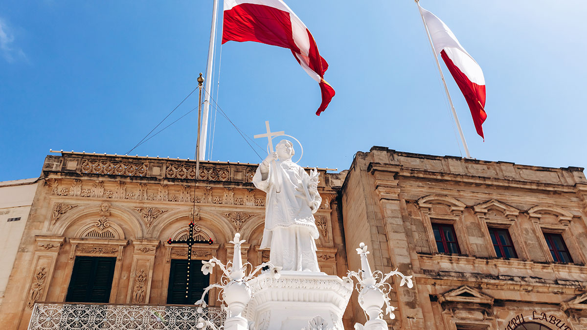 White statue of a saint holding a cross stands against a vivid blue sky, dramatically framed by the bold red-and-white flag of the Knights of St John waving overhead in a traditional Maltese setting